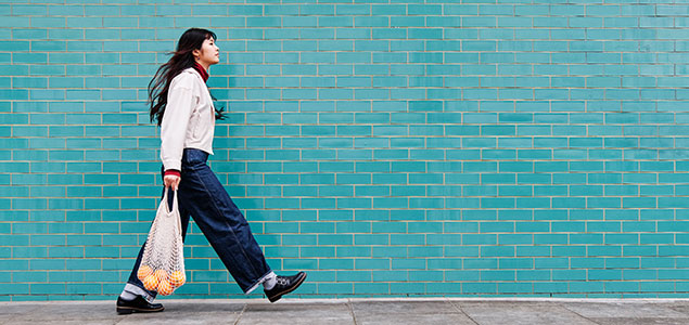 Woman walking in front of a blue-painted wall, holding a bag of oranges